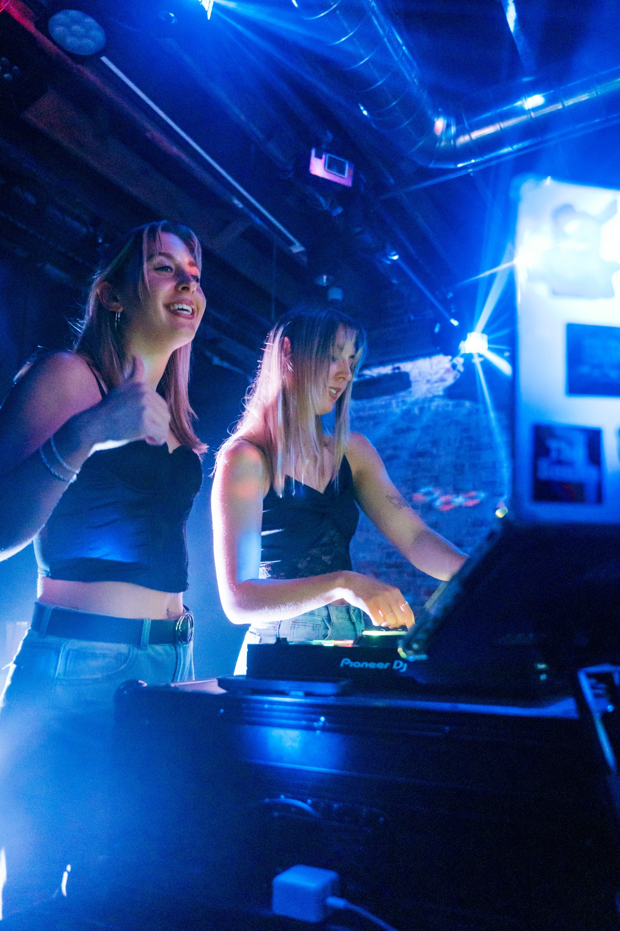 Two women operating DJ equipment in a club setting with blue lighting.