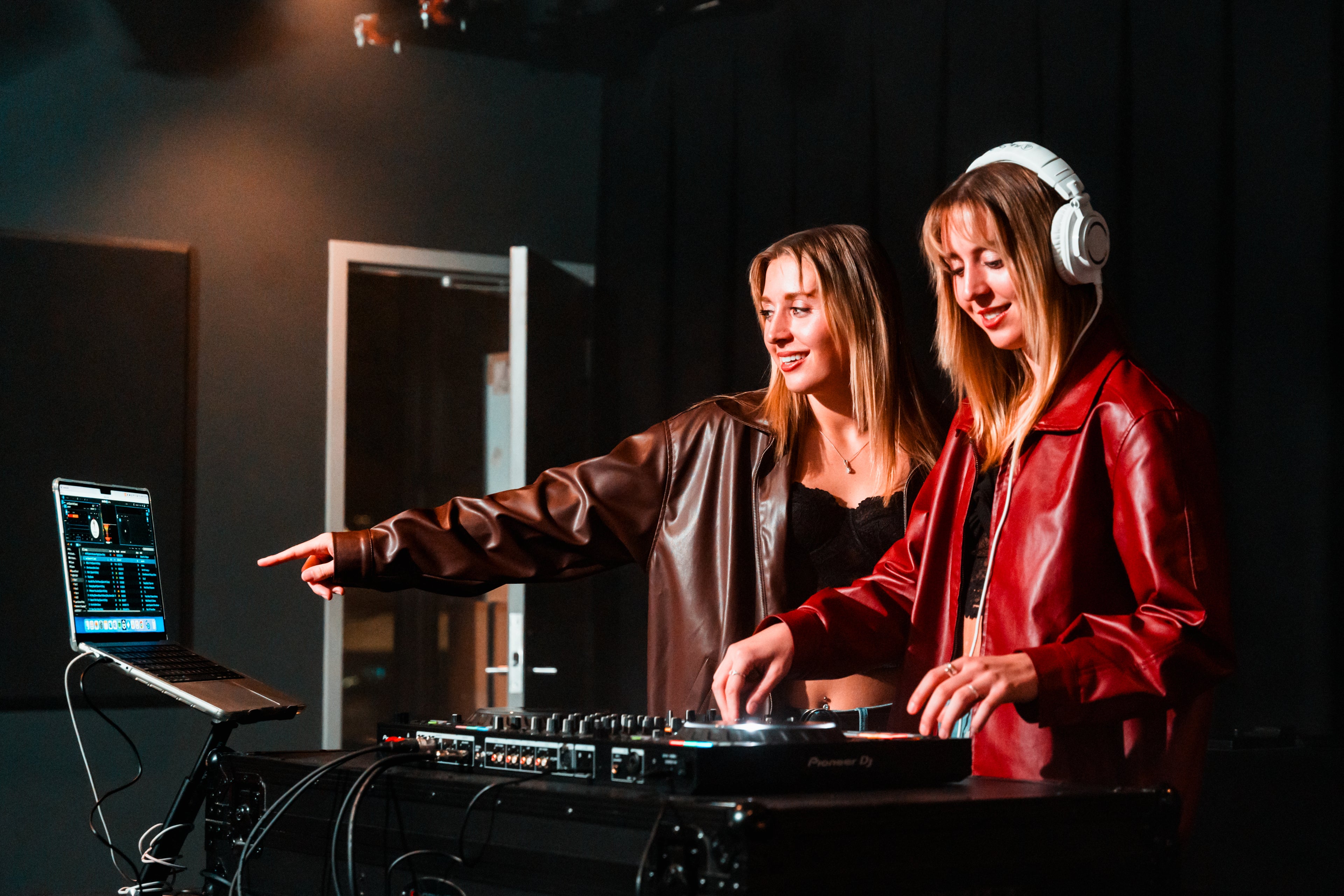 Two women DJing together, one using headphones and pointing at a laptop, both wearing stylish jackets. 