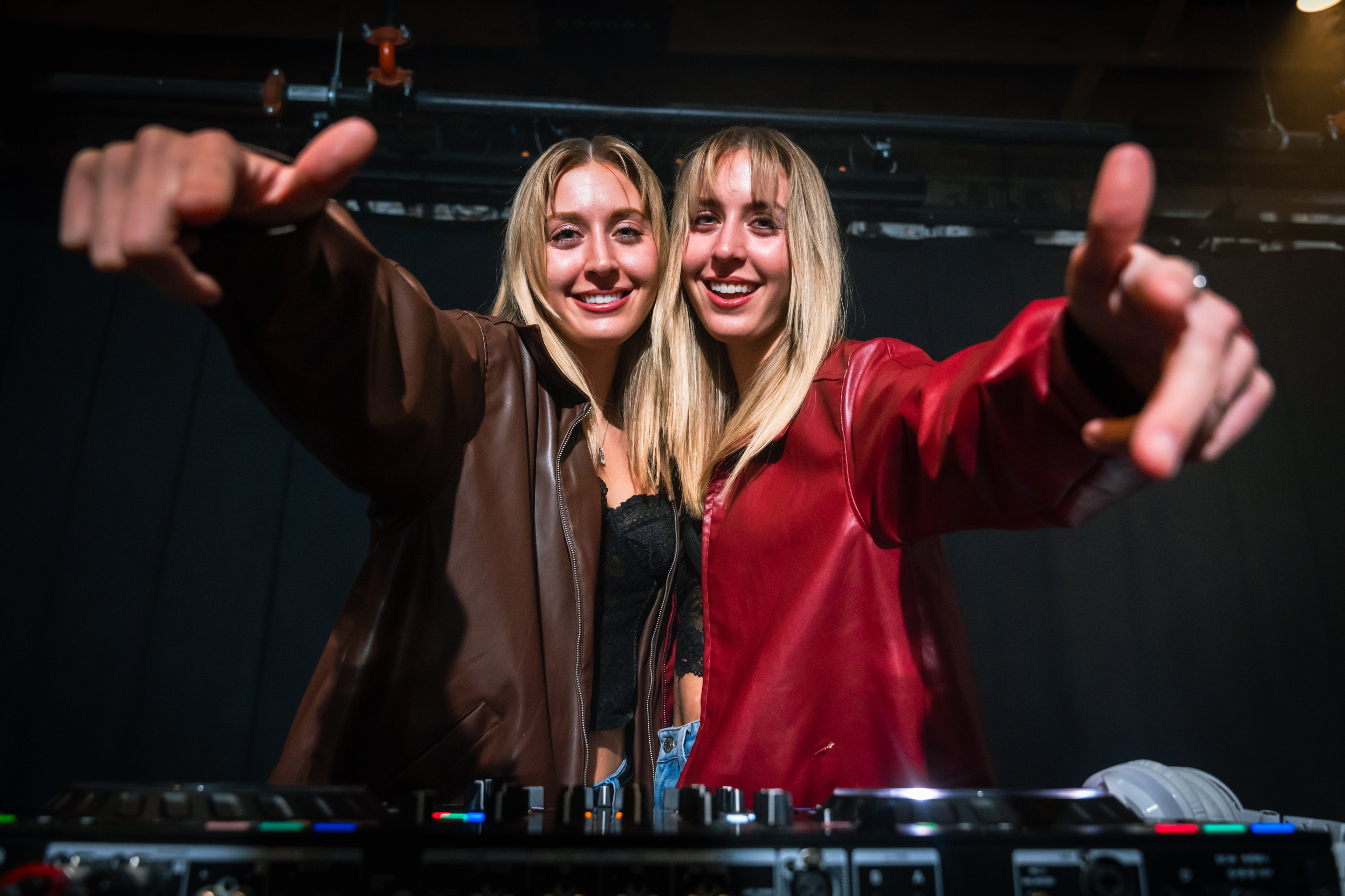 Two women smiling and posing with their arms raised, DJ equipment in front of them. 